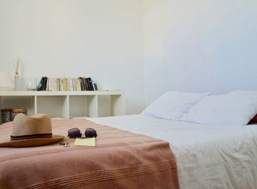 Vue d’une chambre minimaliste avec un lit blanc recouvert d’un plaid rose, posé d’un chapeau de paille, de lunettes de soleil et d’une étagère blanche garnie de livres.