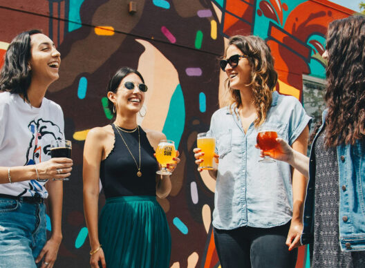 Quatre femmes rieuses tenant des verres de bière artisanale devant un mur coloré orné d'un graffiti multicolore avec l'inscription VEZA SUR sur l'un des verres