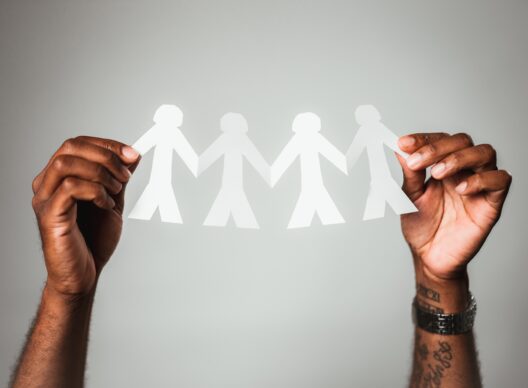 Deux mains de teinte foncée tiennent une guirlande en papier blanc composée de quatre silhouettes humaines reliées par les mains sur fond gris, symbolisant la solidarité, la diversité et le travail d’équipe.