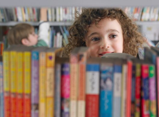 Fille aux cheveux bouclés regardant par-dessus une rangée de livres colorés sur une étagère de bibliothèque, visage souriant et regard curieux.