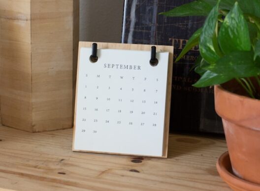 Calendrier de bureau minimaliste sur un support en bois affichant la mention 'September', les abréviations des jours 'S M T W T F S' et les dates numérotées de 1 à 30, posé sur un bureau en bois à côté d’une plante en pot.