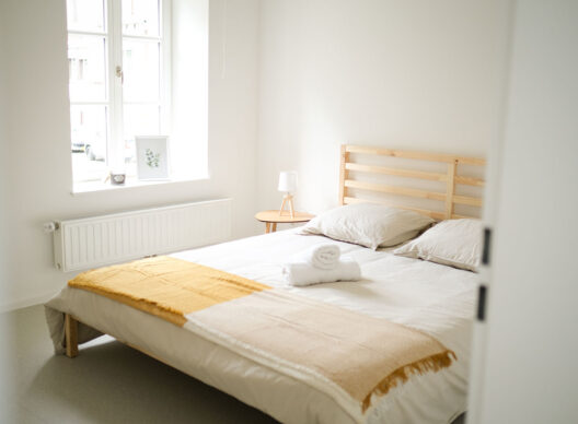 Chambre épurée à la lumière naturelle avec un lit en bois clair, literie blanche, couverture beige et moutarde drapée au pied, serviette roulée, petite table de chevet ronde avec lampe sous une large fenêtre.