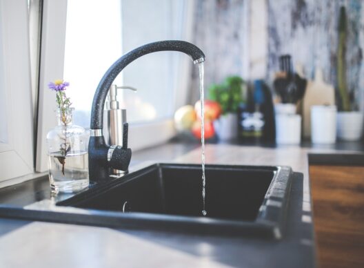 Vue rapprochée d’un évier de cuisine moderne avec robinet noir laissant couler un filet d’eau dans une cuve carrée, bouteille en verre contenant une fleur violette à côté d’une fenêtre, et ustensiles flous en arrière-plan.