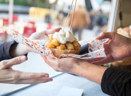 Deux paires de mains s’échangent un bol de pain garni de dés de pommes de terre dorées et croustillantes, nappés d’une crème blanche parsemée de poivre, sur un papier motif graphique