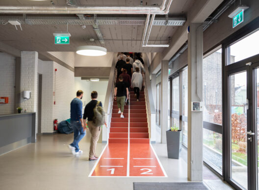Un couloir d’université moderne avec un escalier rouge numéroté 1 et 2 au sol, des étudiants montant, murs en briques blanches, portes vitrées et panneaux de sortie avec flèche verte.