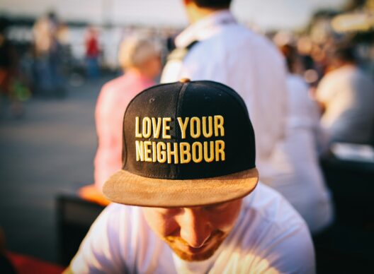 Un homme portant une casquette noire à visière beige brodée en jaune de la phrase « LOVE YOUR NEIGHBOUR », assis devant un arrière-plan urbain flou.