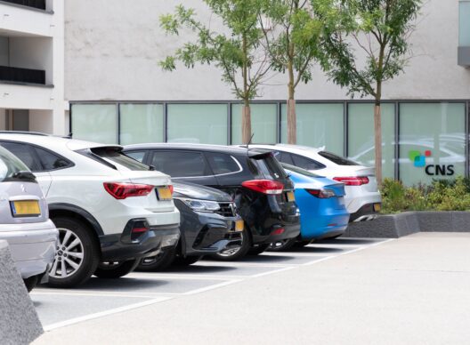 Vue de cinq voitures garées en épi devant la façade vitrée d’un immeuble de bureaux, avec le logo 'CNS' visible sur la vitre. De gauche à droite : une Citroën grise, un SUV blanc, une berline gris foncé, une voiture compacte noire, un véhicule bleu et une berline blanche. Deux petits arbres plantés longent la zone de stationnement.