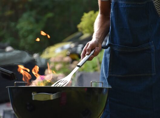 Vue rapprochée d'une personne en tablier en jean utilisant une spatule pour saisir des aliments sur un barbecue au charbon en plein air avec de vives flammes.