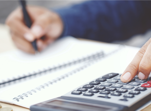Vue rapprochée d’une main droite appuyant sur la touche AC d’une calculatrice, main gauche tenant un stylo pour écrire dans un cahier à spirale, avec une pile de pièces sur un bureau en bois.
