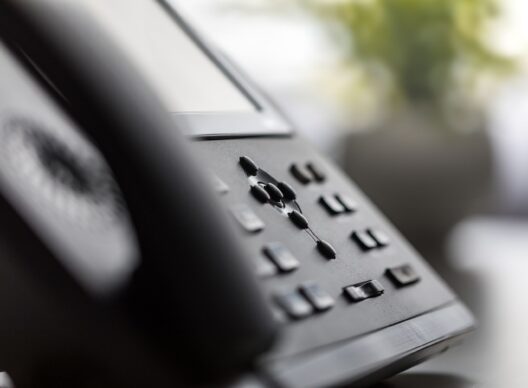 Téléphone de bureau noir posé sur un bureau avec écran numérique, touches fonctionnelles et combiné latéral, arrière-plan flou avec plante verte