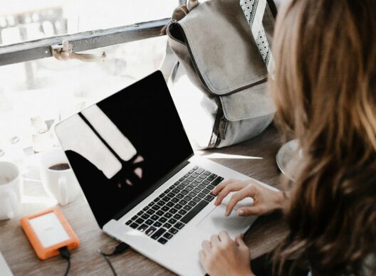 Vue du dessus d’une personne tapant sur un ordinateur portable posé sur un bureau en bois près d’une fenêtre, avec une tasse de café, un disque dur externe orange et un sac à dos gris en arrière-plan.