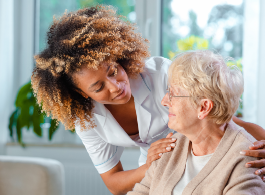 Une infirmière au regard attentionné pose une main rassurante sur l’épaule d’une femme âgée assise près d’une fenêtre, symbolisant l’accompagnement en soins à domicile.