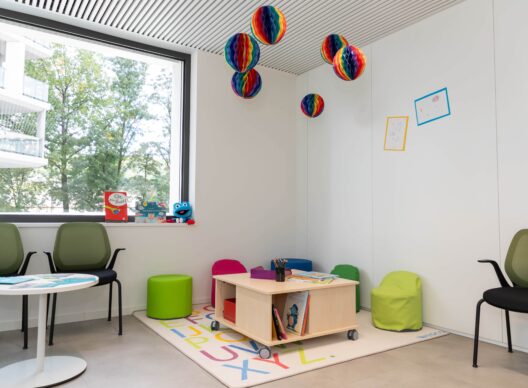 Espace lumineux aménagé pour enfants avec tapis alphabet multicolore, table basse en bois sur roulettes contenant des livres dont le titre « Oje, ein Buch! », peluche grenouille bleue sur le rebord de la fenêtre, petits poufs verts et roses, suspensions en papier arc-en-ciel et dessins d’enfants encadrés au mur