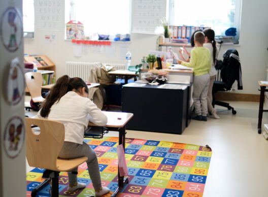 Jeune élève assise sur un tapis coloré numéroté de 1 à 100, écrivant dans son cahier à un pupitre en bois dans une classe lumineuse. À l'arrière-plan, deux enfants échangent près du bureau de l'enseignante. Des affiches murales présentent l'alphabet en majuscules 'A B C D E F G H I J K L M N O P Q R S T U V W X Y Z A O U' et en minuscules 'a b c d e f g h i j k l m n o p q r s t u v w x y z a o u'.