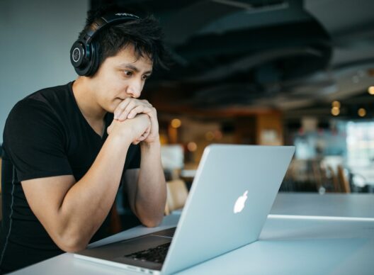 Un jeune homme portant un t-shirt noir et un casque Audio-Technica, assis devant un MacBook sur une table blanche dans un espace de coworking, les mains jointes sous le menton, l'air pensif.