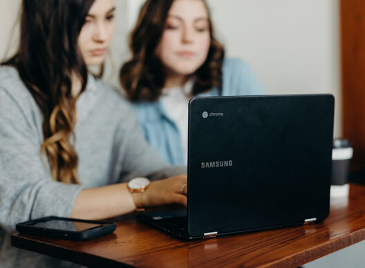 Deux jeunes femmes concentrées sur un ordinateur portable avec les logos "Samsung" et "chrome" posé sur une table en bois, un smartphone et un gobelet de café à emporter à côté.