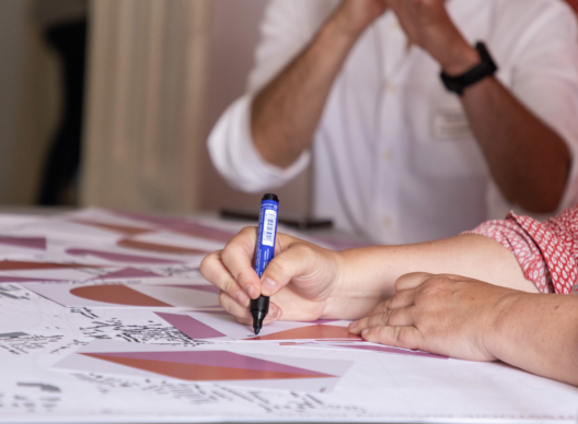 Gros plan sur deux mains dessinant un schéma géométrique en nuances de rose et rouge sur une grande feuille blanche à l’aide d’un feutre bleu, avec une autre personne en chemise blanche en arrière-plan.