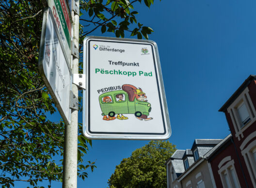 Panneau «Ville de Differdange» affichant «Treffpunkt Pëschkopp Pad» et «PEDIBUS», avec une illustration ludique d’un petit bus vert animé par un écureuil portant un casque et des enfants, sous un ciel bleu.