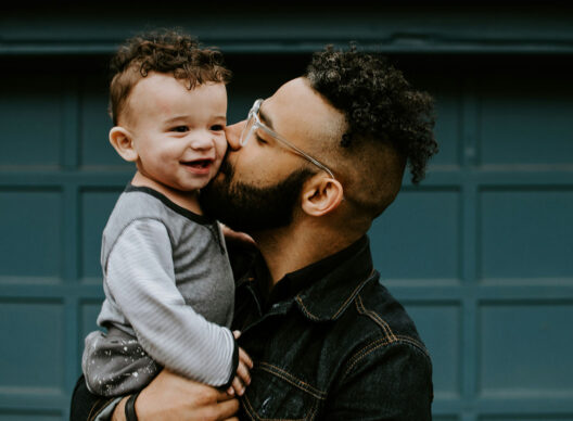 Un père barbu portant des lunettes à monture transparente et une veste en jean tient son bébé bouclé dans ses bras et l’embrasse sur la joue. L’enfant, vêtu d’un pyjama gris, sourit devant un portail bleu.