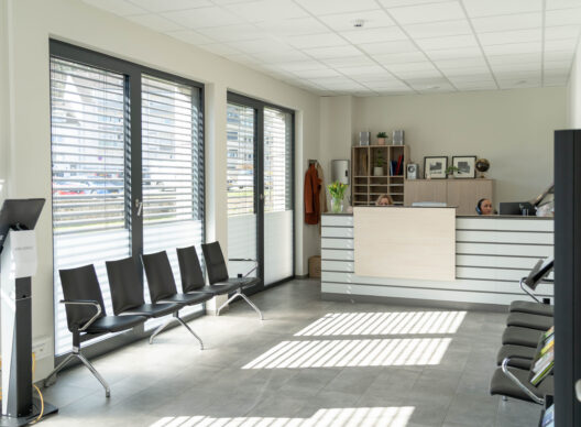 Salle d’attente médicale spacieuse et lumineuse avec chaises noires alignées, comptoir de réception en bois clair avec deux hôtesses partiellement visibles, grandes baies vitrées équipées de stores vénitiens projetant des ombres rayées, vase de tulipes jaunes et pupitre à écran marqué "HORS SERVICE"