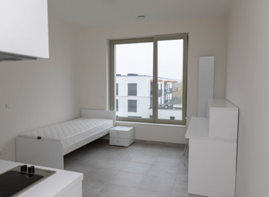 Studio blanc lumineux avec lit simple, chevet, kitchenette à induction, bureau épuré et grande fenêtre donnant sur des immeubles résidentiels modernes.