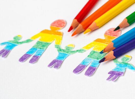Découpe papier représentant une famille de quatre silhouettes (deux parents et deux enfants) unies par les mains, colorées de bandes arc-en-ciel sur fond blanc, accompagnée de crayons de couleur assortis.